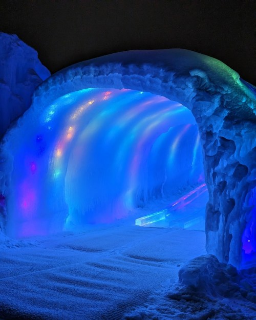 A glowing ice tunnel illuminated with colorful lights, surrounded by snow.