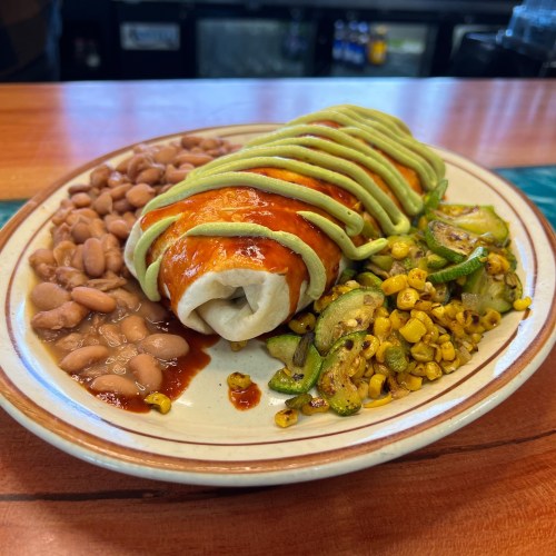 A plate featuring a burrito topped with green sauce, served with pinto beans and sautéed corn and zucchini.