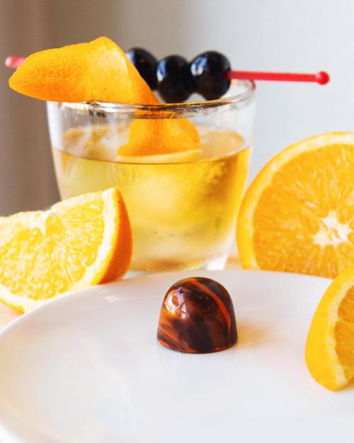 A glass of cocktail garnished with orange peel and cherries, alongside chocolate truffle and orange slices on a plate.