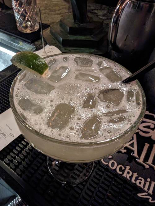 A close-up of a frothy cocktail in a large glass, garnished with a lime wedge and filled with ice.