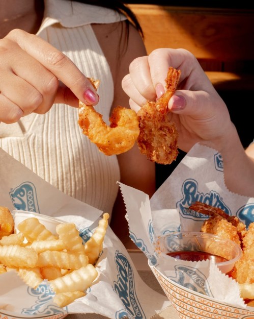Two hands hold crispy shrimp above a basket of fries, with a dipping sauce on the side.