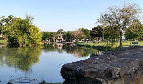 A serene park scene featuring a calm pond, lush greenery, and a stone wall in the foreground under a clear blue sky.