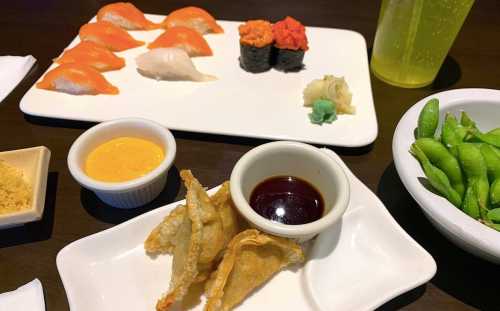 A plate of sushi, including nigiri and rolls, with dipping sauces, edamame, and crispy dumplings on the side.