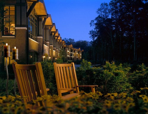 Two wooden rocking chairs sit in a garden, illuminated by soft lights, with a modern building in the background at dusk.