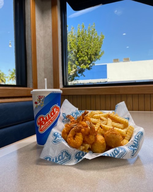 A plate of crispy shrimp and fries next to a large drink, with a sunny outdoor view through the window.