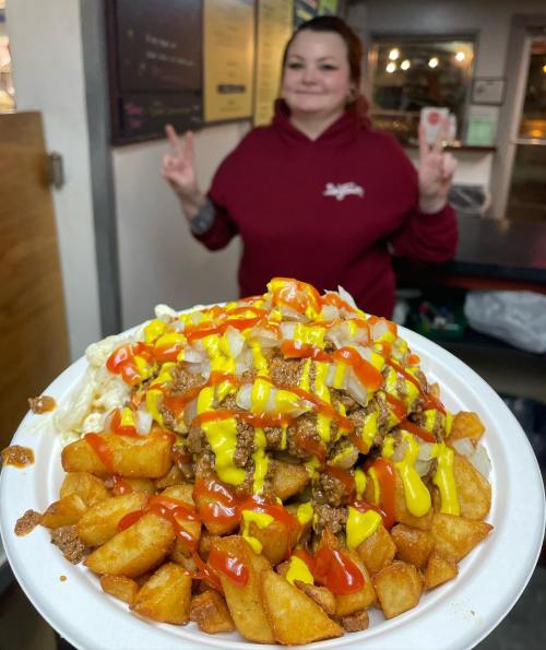A plate of loaded fries topped with cheese, onions, and condiments, with a person giving a peace sign in the background.