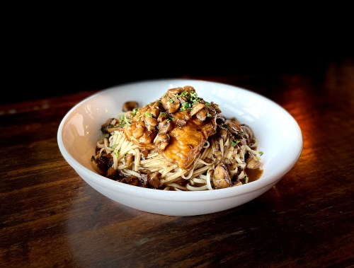 A bowl of noodles topped with chicken and mushrooms, garnished with green herbs, on a wooden table.