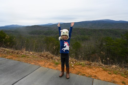 A joyful child in a unicorn sweater and hat stands with arms raised, overlooking a scenic mountain view.