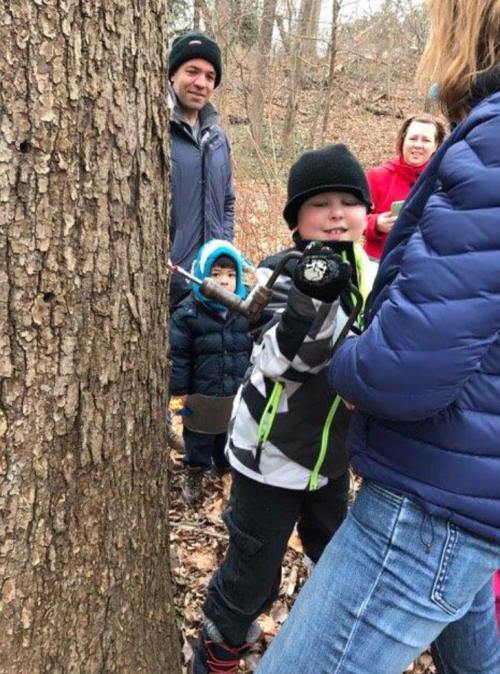 A child in a black and white jacket holds a stick, while adults and another child watch in a wooded area.