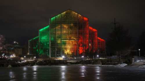 A brightly lit greenhouse at night, illuminated in red and green lights, reflecting on a frozen surface nearby.