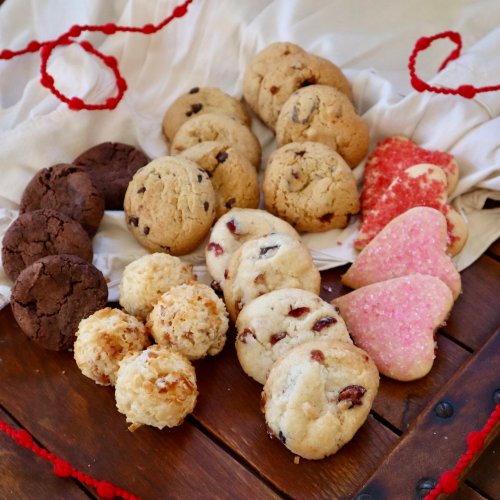A variety of cookies arranged on a white cloth, including chocolate, sugar, and coconut cookies, with red decorations.