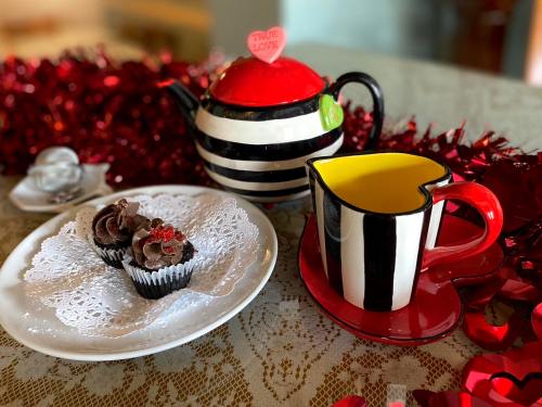 A striped teapot and cup with a plate of chocolate cupcakes, decorated with red hearts, on a lace tablecloth.