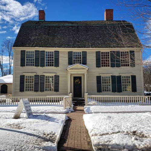 Historic two-story house with a steep roof, green shutters, and a snow-covered yard under a bright blue sky.