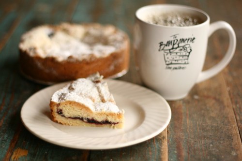 A slice of cake dusted with powdered sugar on a plate, next to a coffee mug and a whole cake in the background.