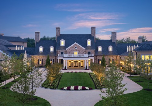 A grand stone mansion with a landscaped front yard, illuminated by soft lights at dusk.