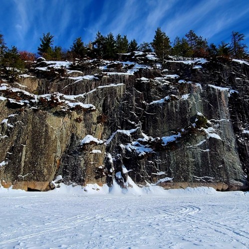 Snow-covered rocky cliff with trees on top, under a blue sky with wispy clouds.