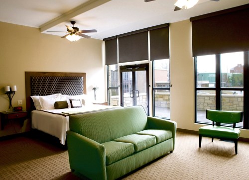 A spacious hotel room featuring a bed, green sofa, chair, and large windows with natural light.