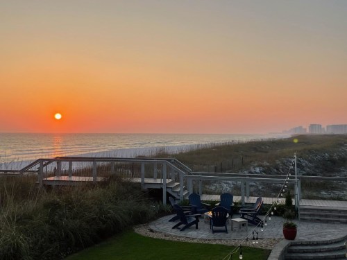Sunset over a beach with a wooden deck, chairs, and distant buildings along the shoreline. Soft colors in the sky.