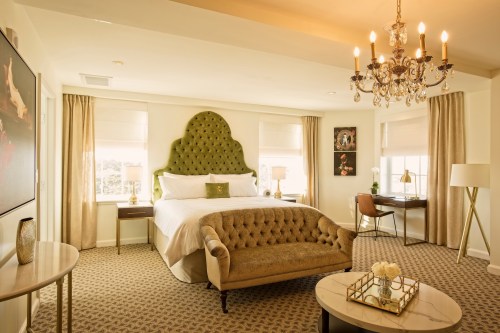 A stylish bedroom featuring a green tufted headboard, a chandelier, and elegant furnishings with natural light.