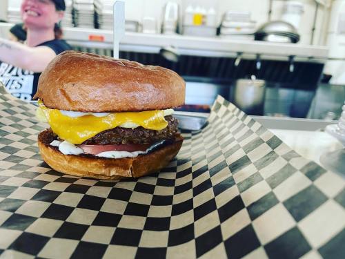 A close-up of a burger with cheese and a fried egg on a checkered paper plate, with a smiling staff member in the background.