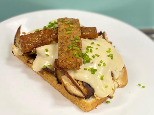 A slice of toasted bread topped with melted cheese, tempeh strips, and chopped chives on a white plate.