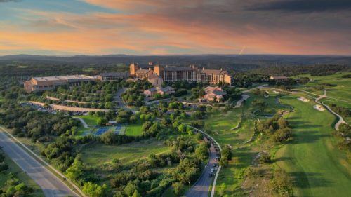 Aerial view of a resort surrounded by lush greenery and rolling hills under a colorful sunset sky.
