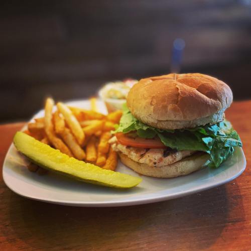 A plate with a grilled chicken sandwich, lettuce, tomato, a pickle, fries, and a small serving of coleslaw.