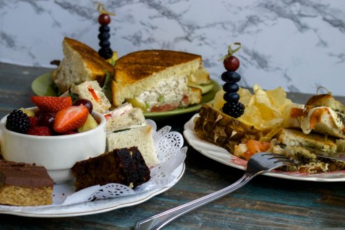 A variety of sandwiches, fruit salad, and desserts on plates, with a rustic wooden table and marble background.