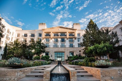 A modern hotel with a stone facade, surrounded by lush greenery and a water feature, under a blue sky with clouds.
