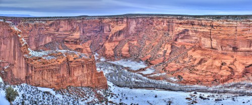 A panoramic view of a canyon with red rock formations, snow-covered ground, and a winding river below.