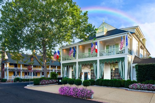 A charming building with flags, surrounded by greenery and flowers, under a rainbow in a clear blue sky.