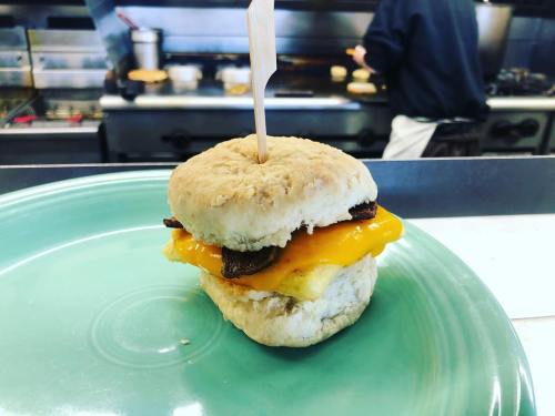 A biscuit sandwich with cheese, bacon, and egg, served on a green plate with a toothpick. Kitchen in the background.