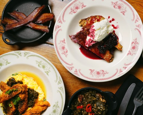 A table spread featuring French toast with berries and whipped cream, fried chicken, greens, and crispy bacon.
