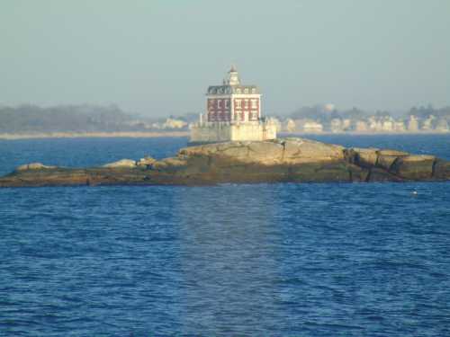 A small, historic lighthouse stands on a rocky island, surrounded by calm blue waters and distant shoreline.