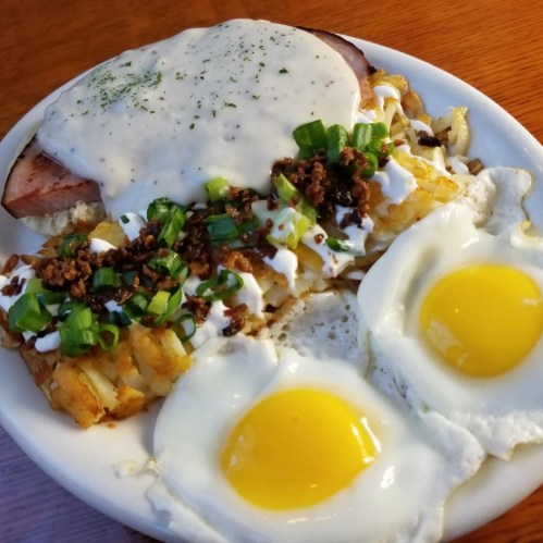A plate of breakfast featuring two sunny-side-up eggs, hash browns topped with green onions, and sausage gravy over ham.