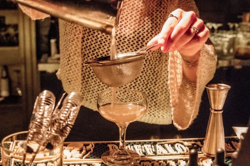 A bartender pours a cocktail through a strainer into a glass, with bar tools and ingredients visible in the background.