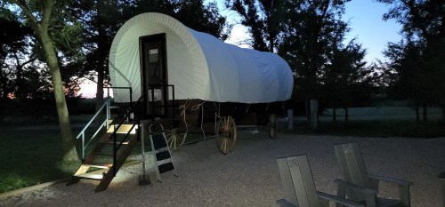 A covered wagon on a gravel area, illuminated at night, surrounded by trees and a few chairs nearby.