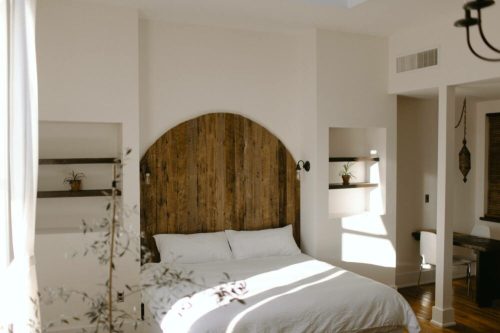Cozy bedroom with a wooden headboard, white bedding, and natural light streaming through large windows.
