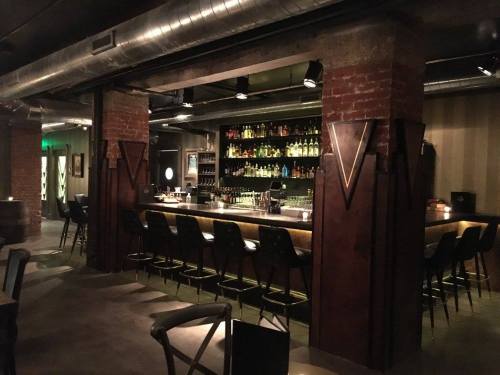 A dimly lit bar with a sleek counter, high stools, and shelves lined with various bottles against a brick wall.