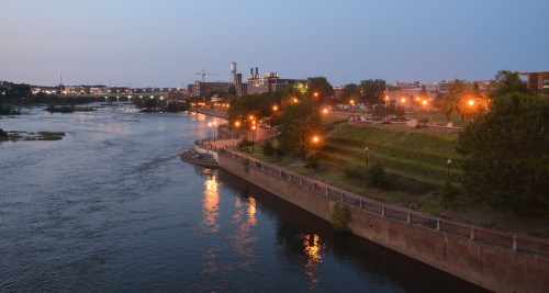 A serene river scene at dusk, with city lights reflecting on the water and a park along the shore.