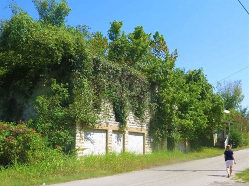 A person walks along a road beside an overgrown, ivy-covered building under a clear blue sky.