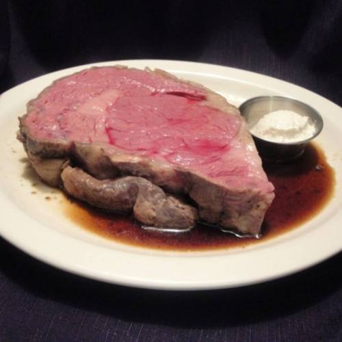 A large, juicy slice of prime rib served on a plate with a small bowl of horseradish sauce and a dark gravy.