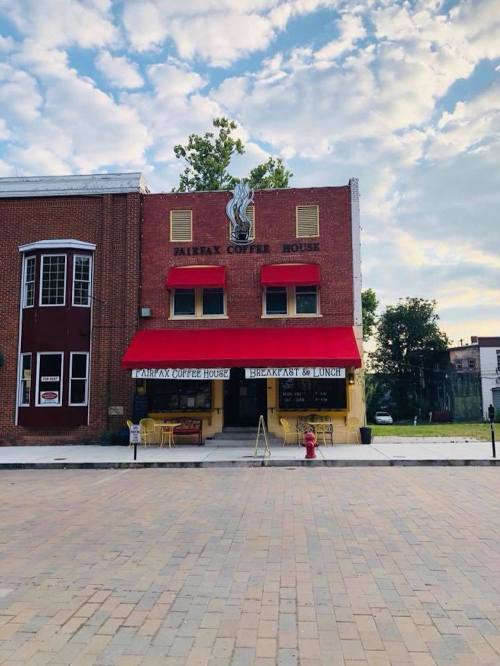 A brick coffee house with red awnings, outdoor seating, and a blue sky with clouds in the background.
