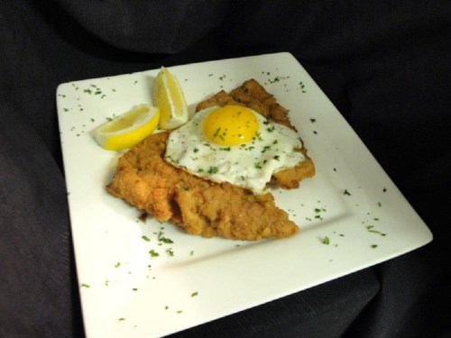 A plate of fried fish topped with a sunny-side-up egg, garnished with parsley and served with lemon wedges.