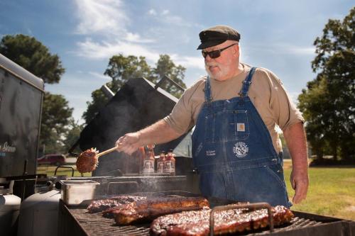 A man in overalls grills ribs outdoors, surrounded by smoke and barbecue sauce under a clear blue sky.