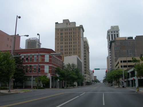 A quiet city street lined with tall buildings and traffic lights under a cloudy sky.