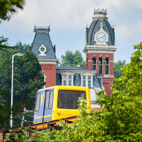 A yellow monorail passes by a historic clock tower building in a green, tree-lined area.