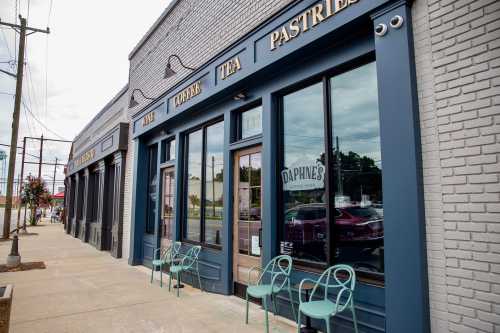 Exterior of a café with large windows, blue trim, and green chairs outside, featuring signs for coffee, tea, and pastries.