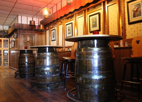 Interior of a rustic bar with wooden floors, barrel tables, and framed artwork on the walls. Warm lighting creates a cozy atmosphere.