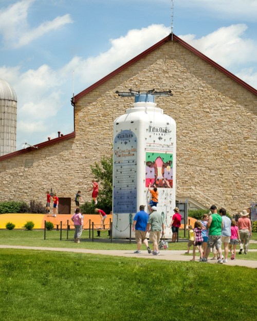 A colorful silo with climbing features, surrounded by people enjoying a sunny day at a farm-themed attraction.